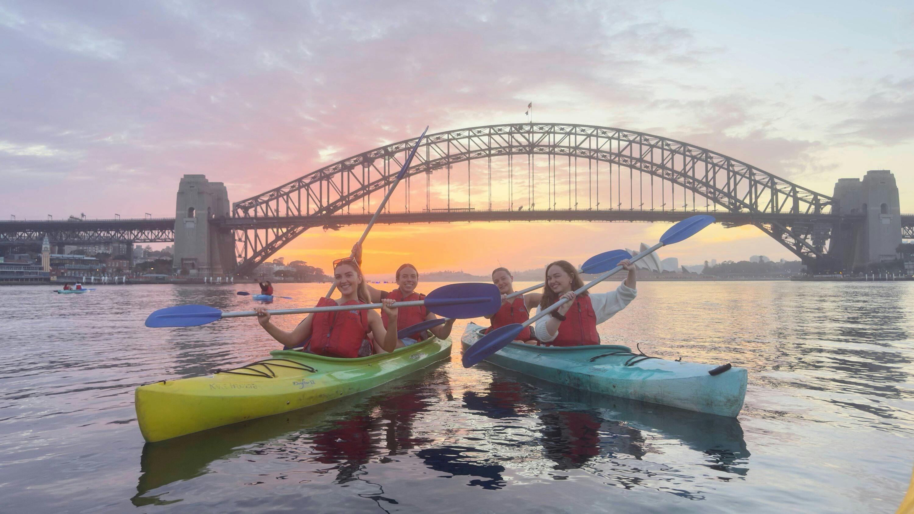 一群皮划艇愛好者在雪梨港的日出中劃行，背後是海港大橋和城市天際線。