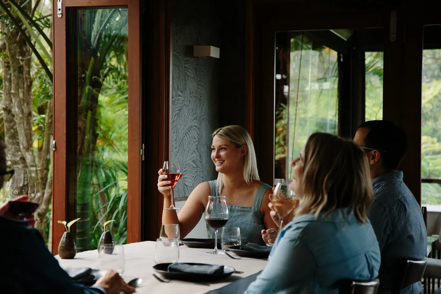 A group of people dining in a restaurant holding glasses of wine, surrounded by rainforest views