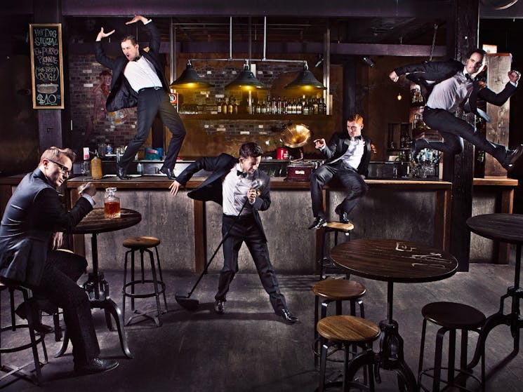 Tap ensemble scattered around a dimly lit bar setting dressed in tuxedos