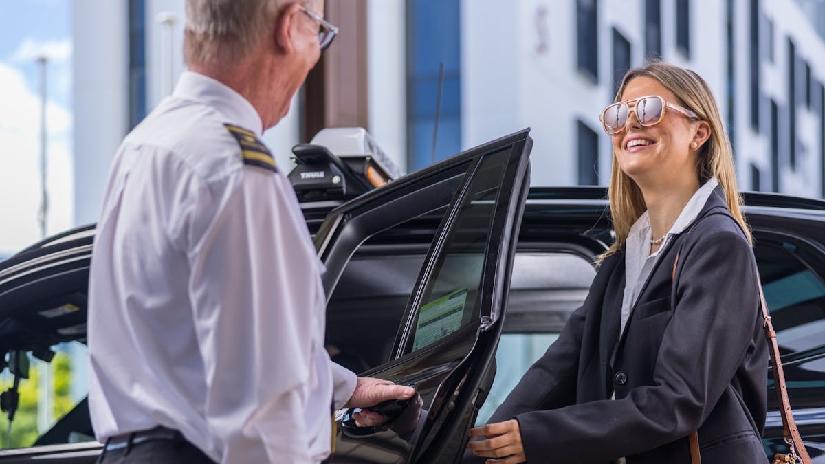 Driver holding open the door for a young female passenger who is dressed professionally and smiling