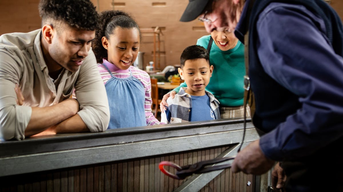 A family watches a blacksmith who is holding heated metal in tongs. A family watches a blacksmith who is holding heated metal in tongs.