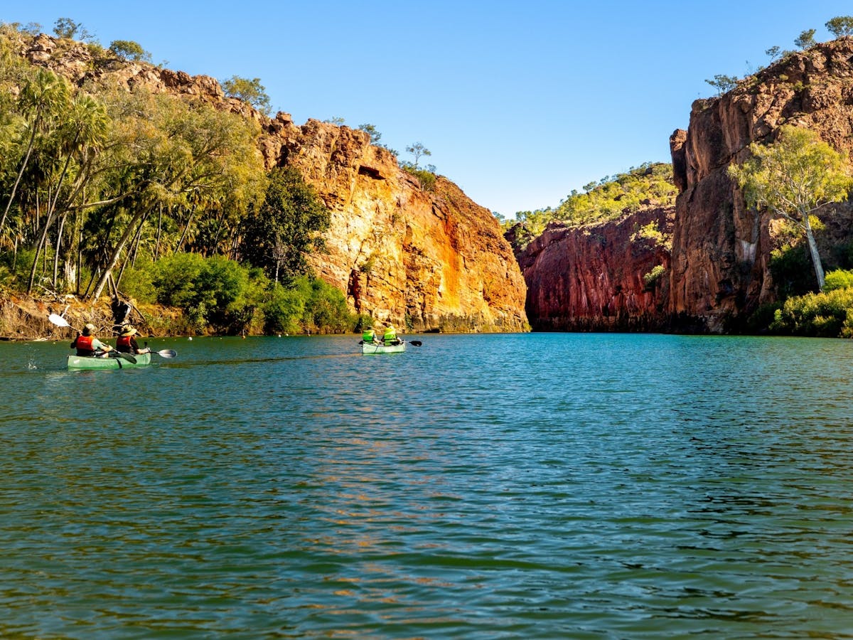 Two canoes on a calm river beneath steep red cliffs and green vegetation under a clear blue sky.