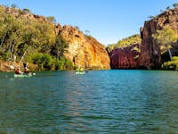 Two canoes on a calm river beneath steep red cliffs and green vegetation under a clear blue sky.