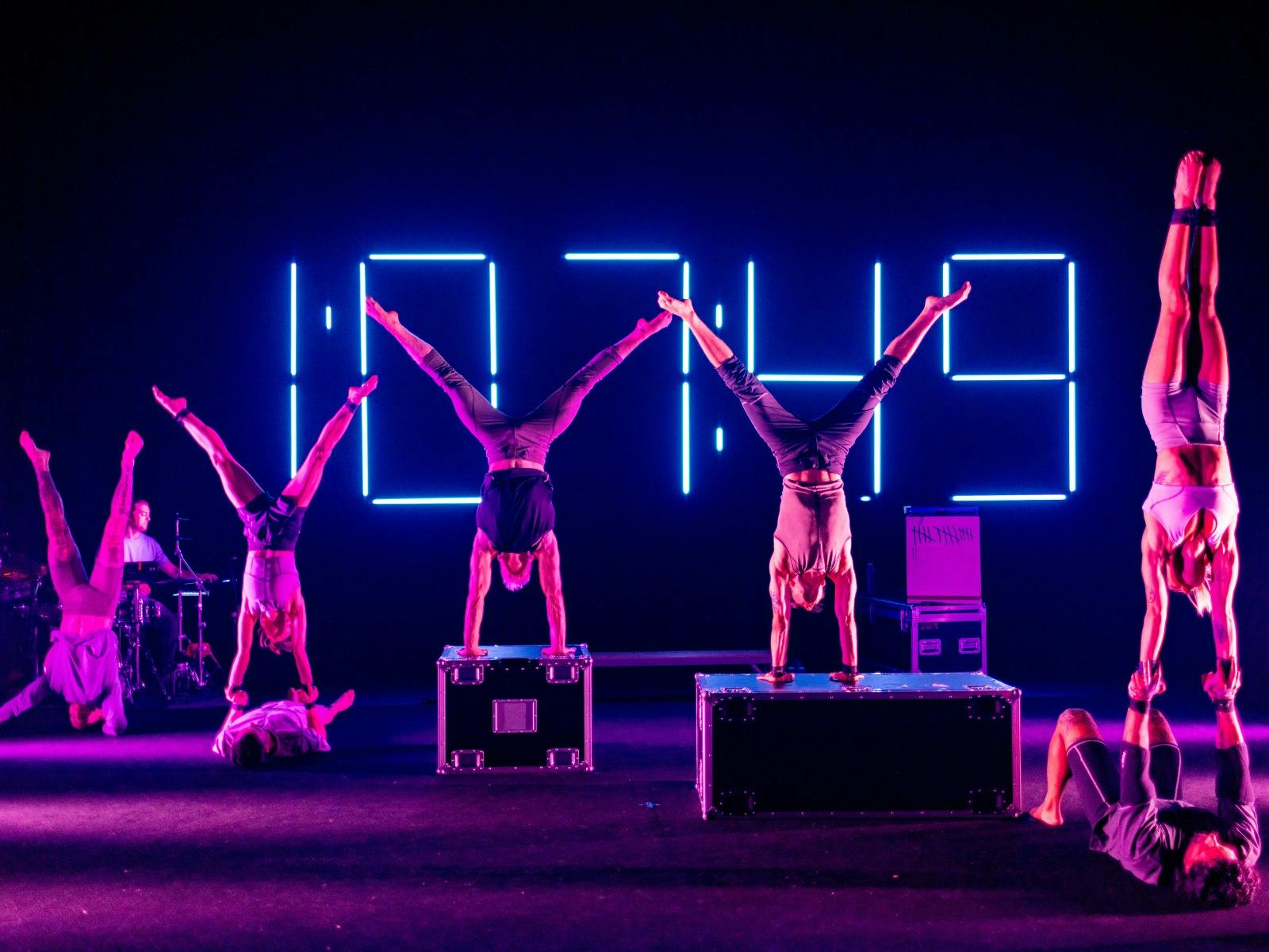 A group of acrobats performing stunts lit by coloured lighting with a neon clock in the background.