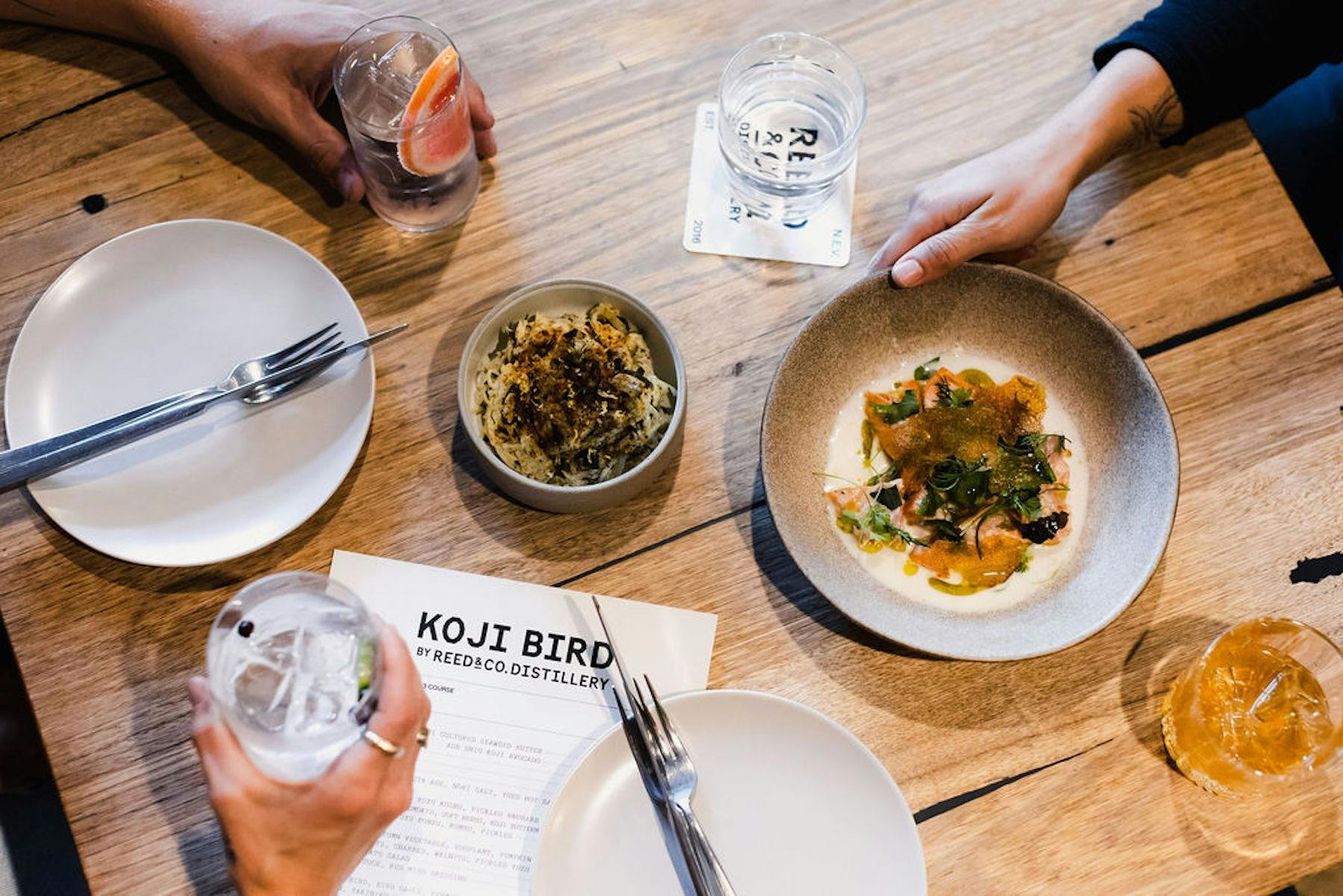 Food and drinks lay on a table in a restaurant, with hands holding some of them