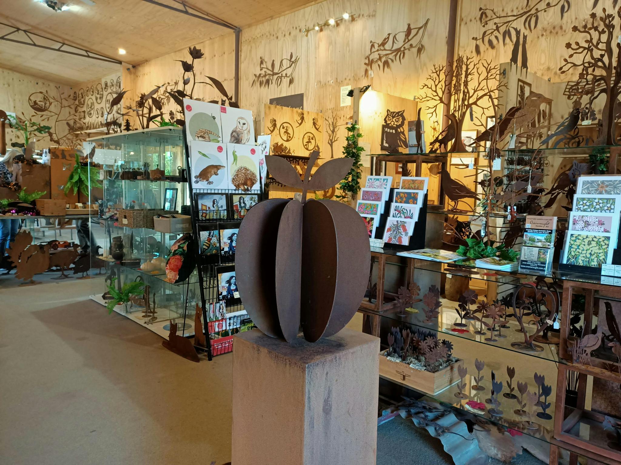 A large metal apple sites on a plinth, surrounded by other metal art and some floral prints & cards.
