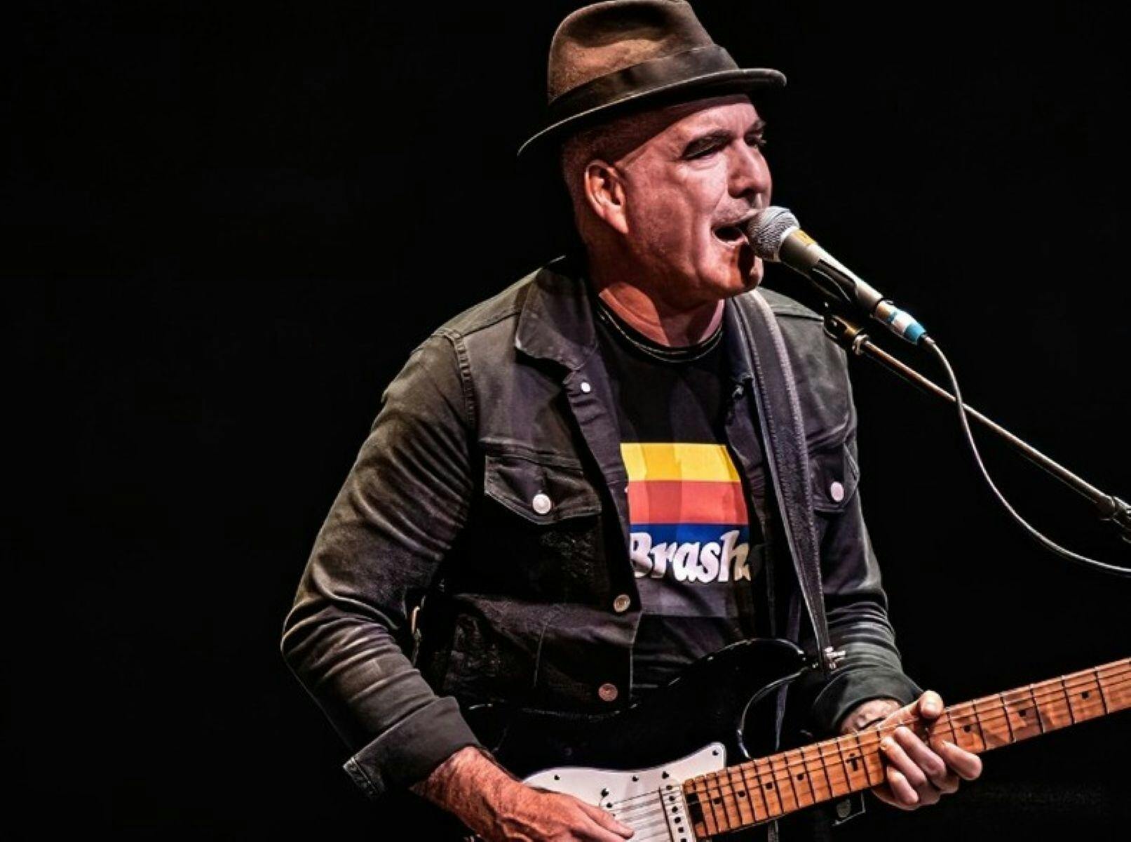 Man wearing fedora playing guitar and singing in to microphone