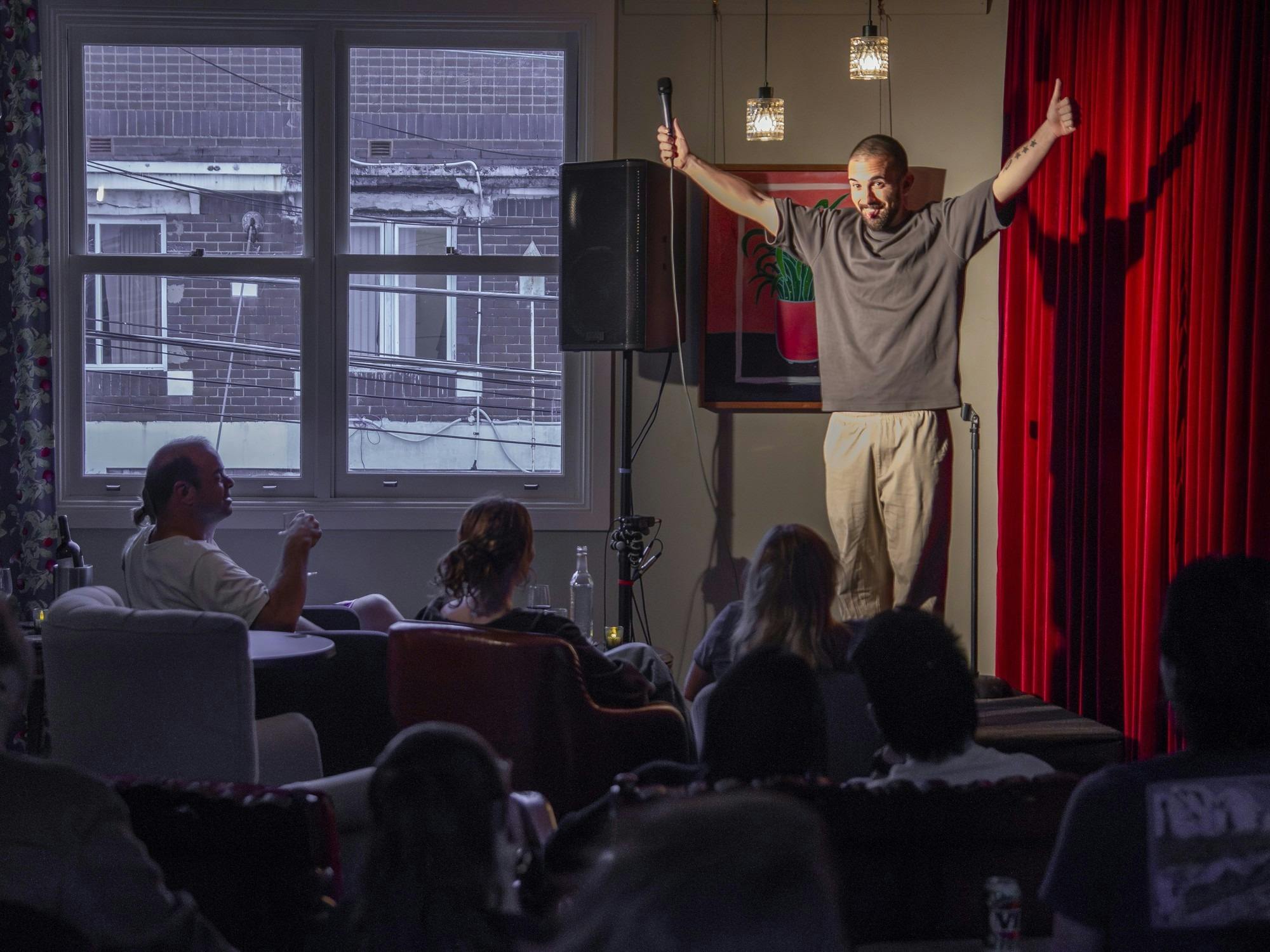 Stand-up comedian performing in front of a red curtain to an audience on leather couches