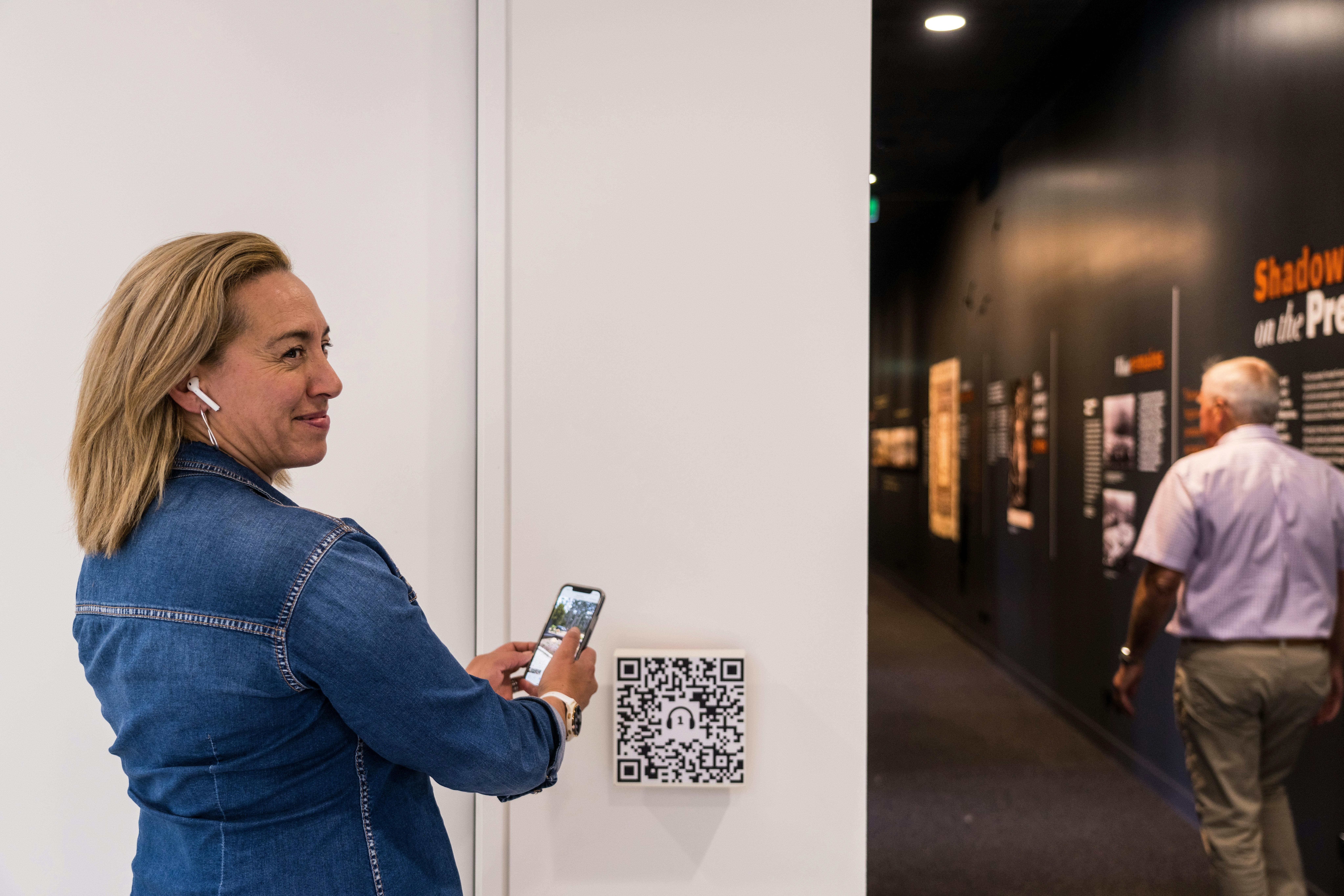 A lady wearing a headphone standing in front of the gallery scanning a QR code for Audio Tour