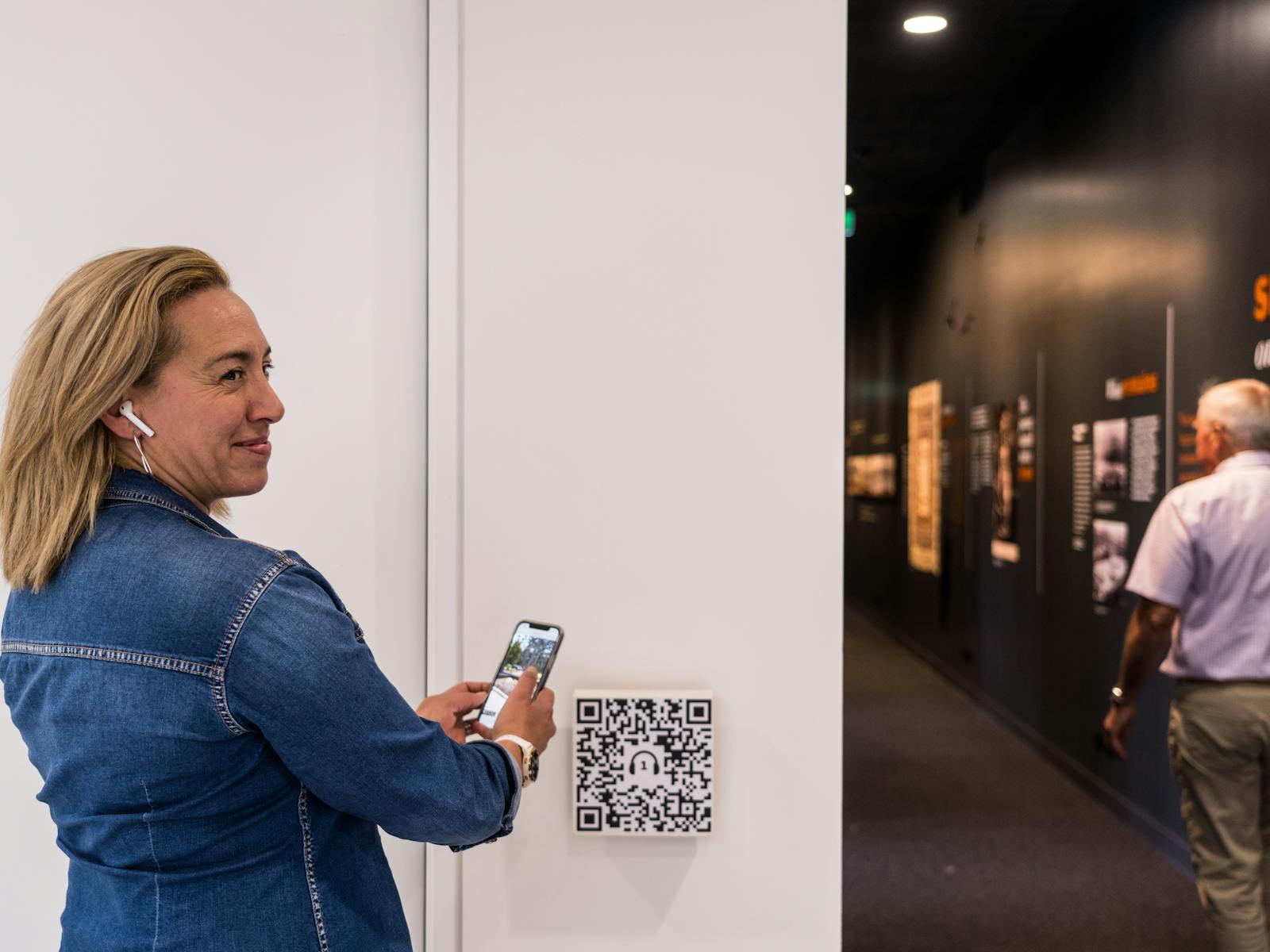 A lady wearing a headphone standing in front of the gallery scanning a QR code for Audio Tour