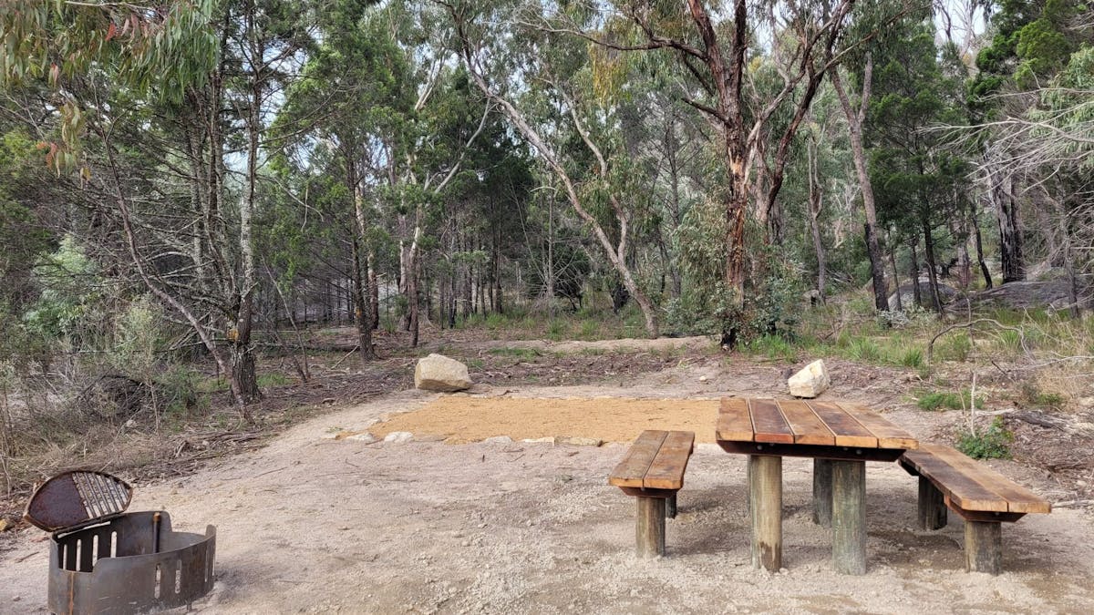 A flat, dirt camp site, featuring a fire pit and picnic table.