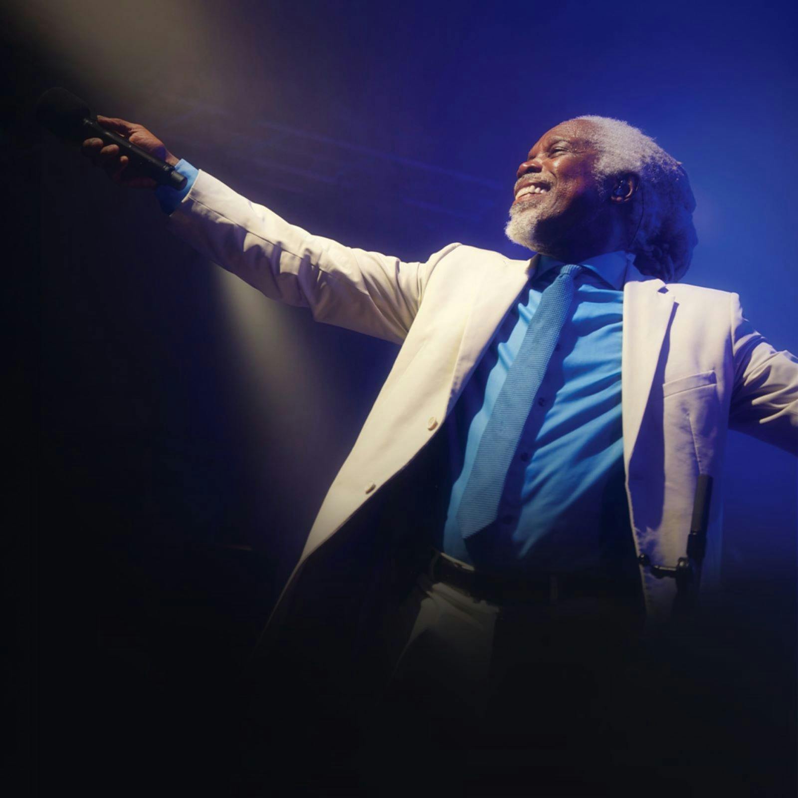 Photo of Billy Ocean in a white suit jacket and blue shirt and tie with his arm out