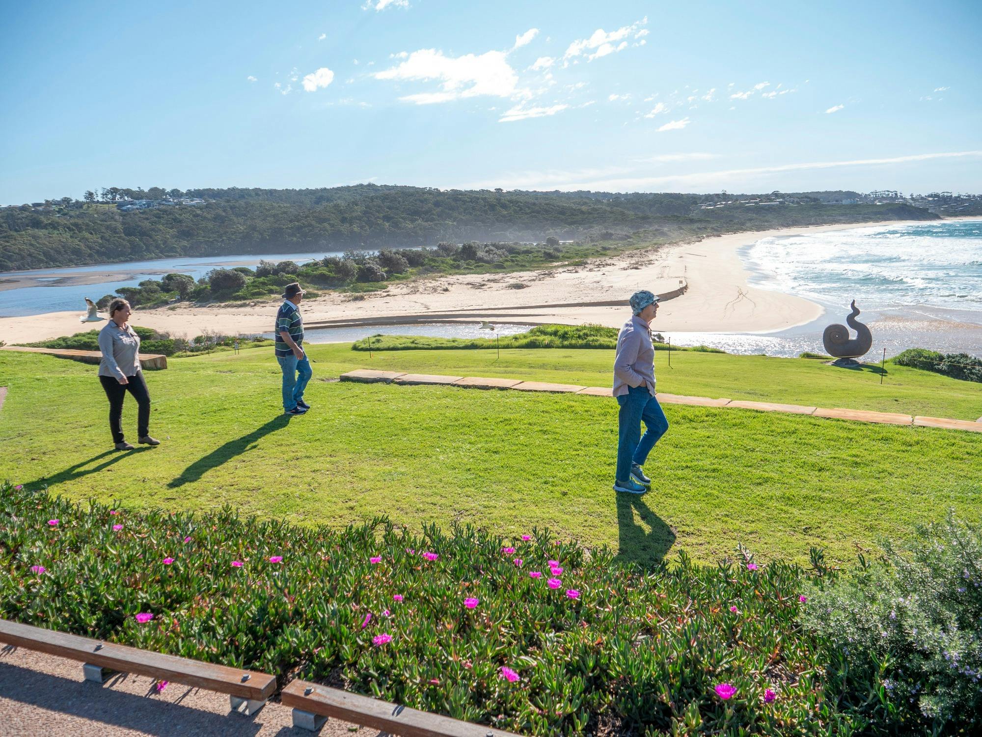 Guests walking on a grassy headland at Short Point, Merimbula Peninsula, with Short Point Beach visi