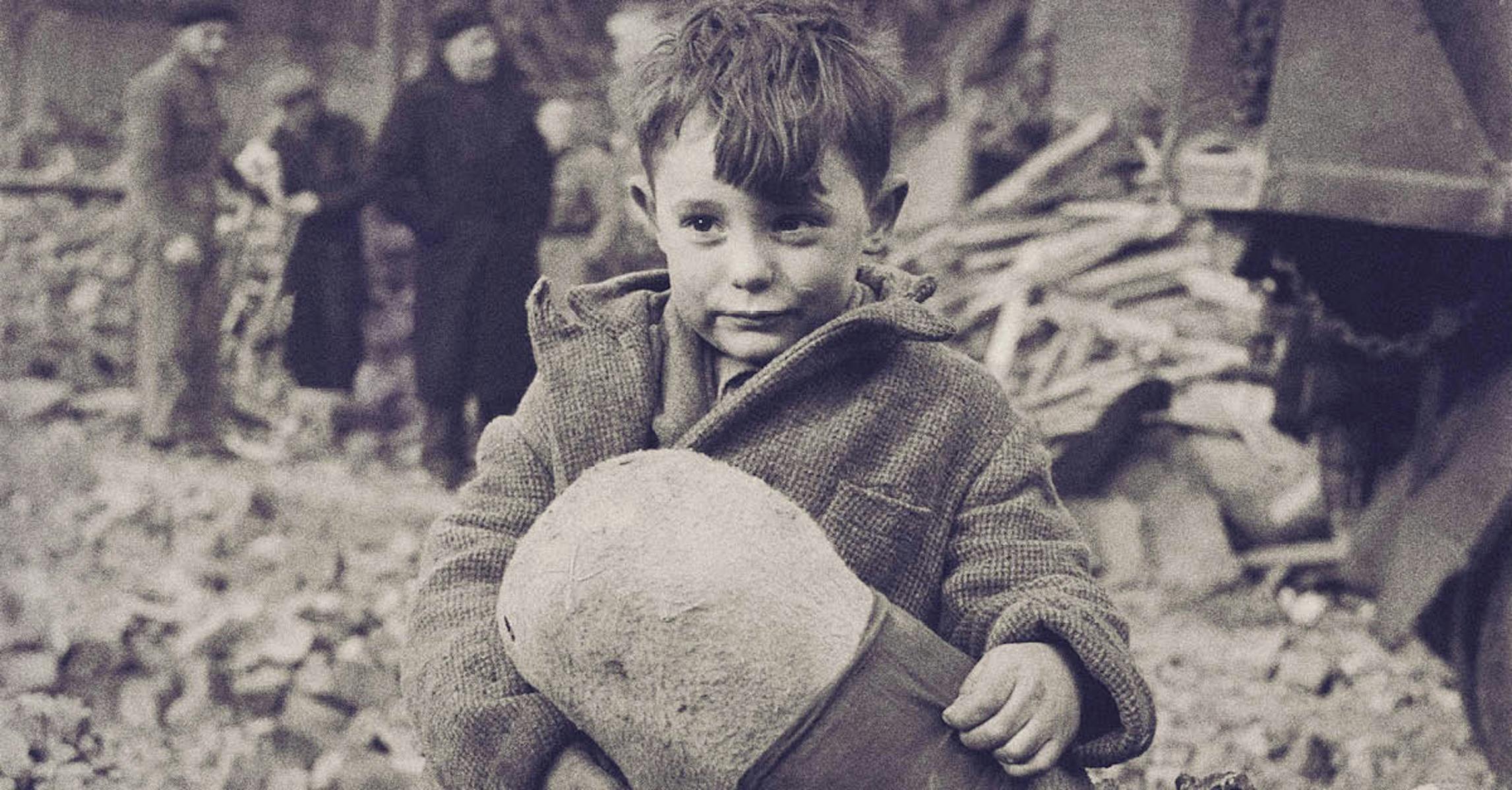 A young boy sits on rubble holding a stuffed toy, with damaged buildings and people in background