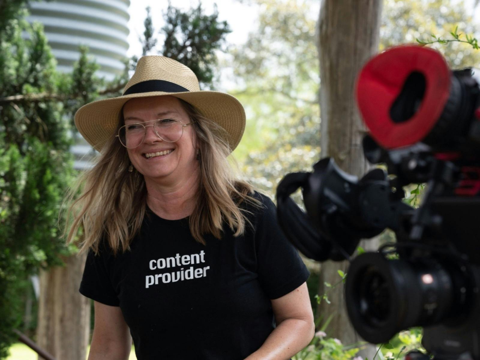 A blonde woman in a beige hat wears a black t-shirt. She stands smiling next to a camera