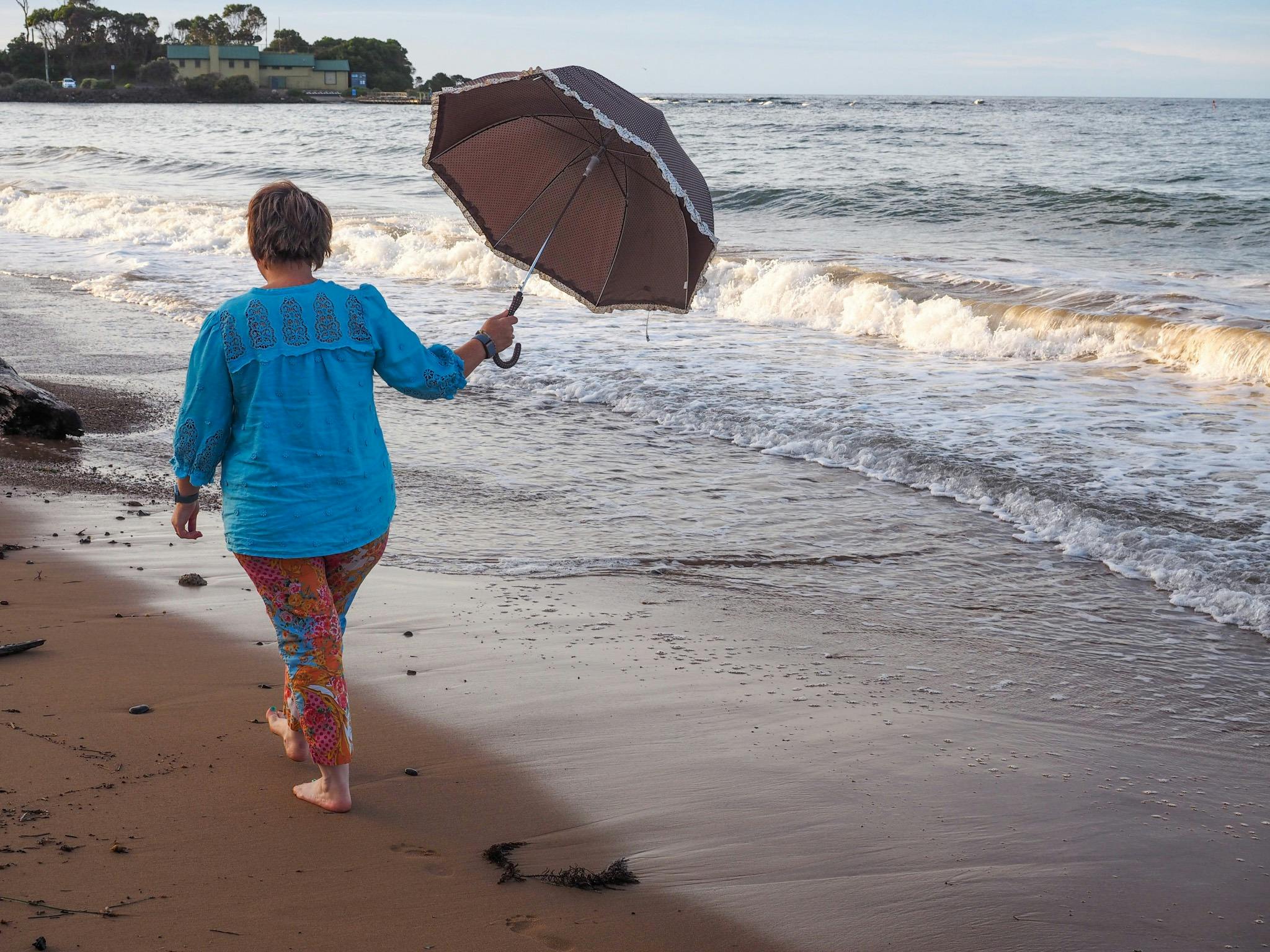 Colourful woman walking barefoot on Penguin Beach, TAS with an umbrella open,  ready for seamist!