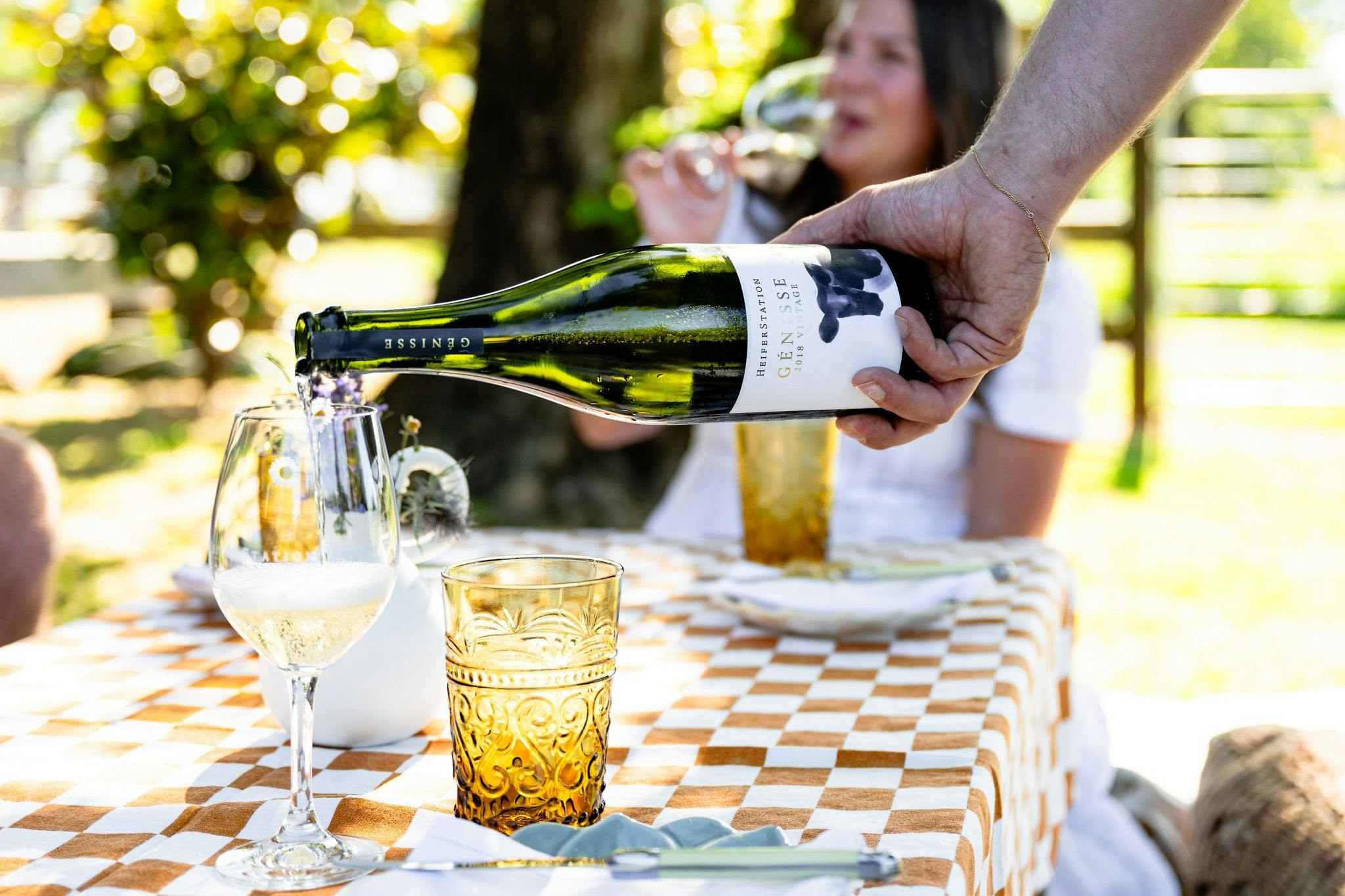 A hand pours a glass of sparkling wine into a glass in an outdoor picnic setting.