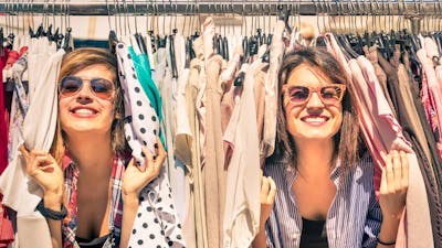two women smiling peeking through a rack of clothes at an outdoor market