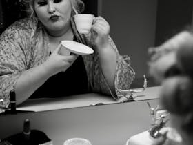 Charlee Watt with hair in curlers holding a coffee cup and gazing into a mirror, contemplating.