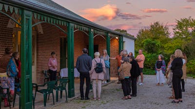 People gather on the verandah of a historic red brick house. The sun sets above.