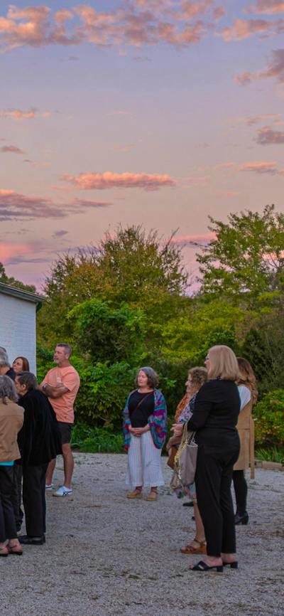 People gather on the verandah of a historic red brick house. The sun sets above.
