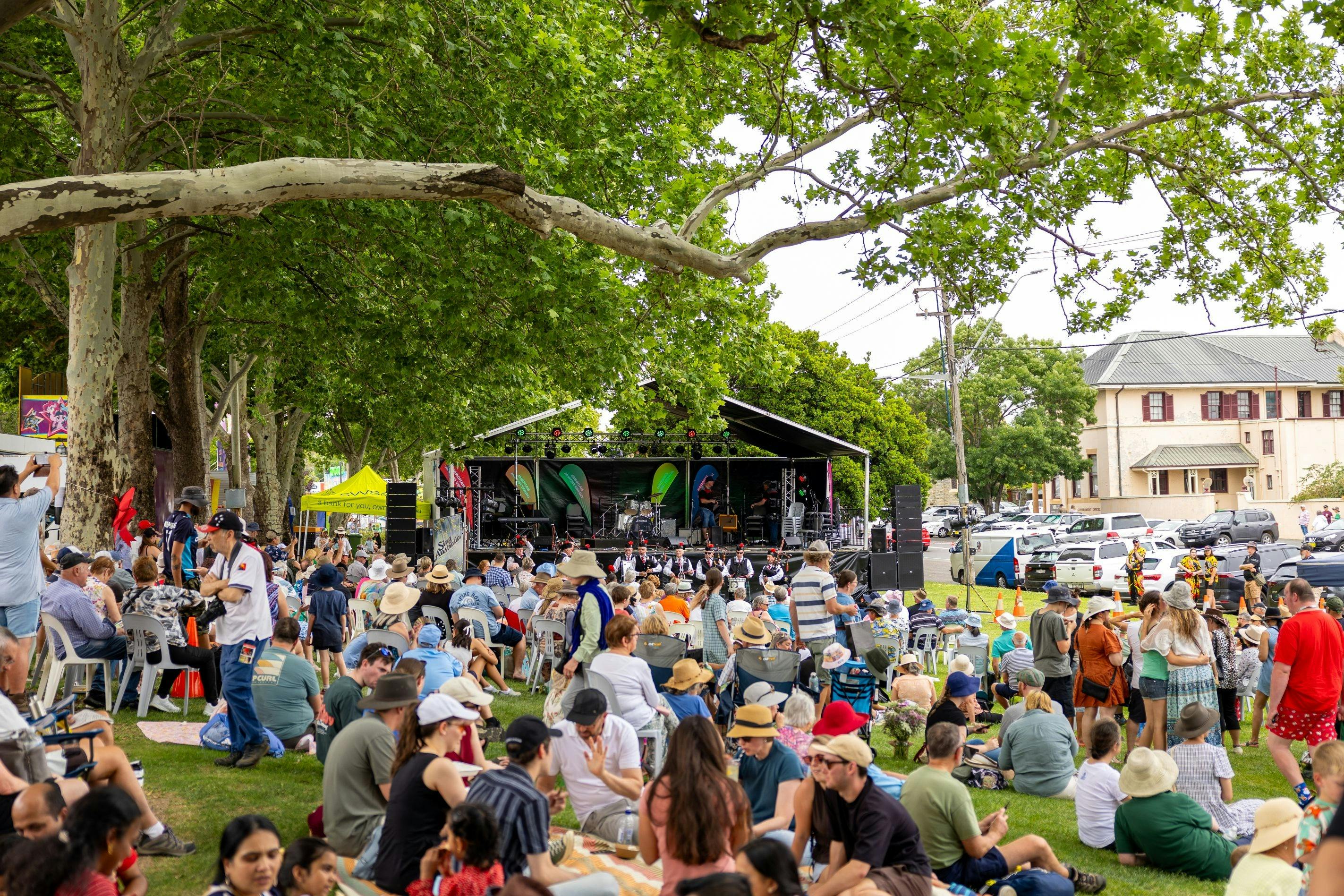 Shot of the main stage from far away, a crowd of people relaxing in between under green trees.