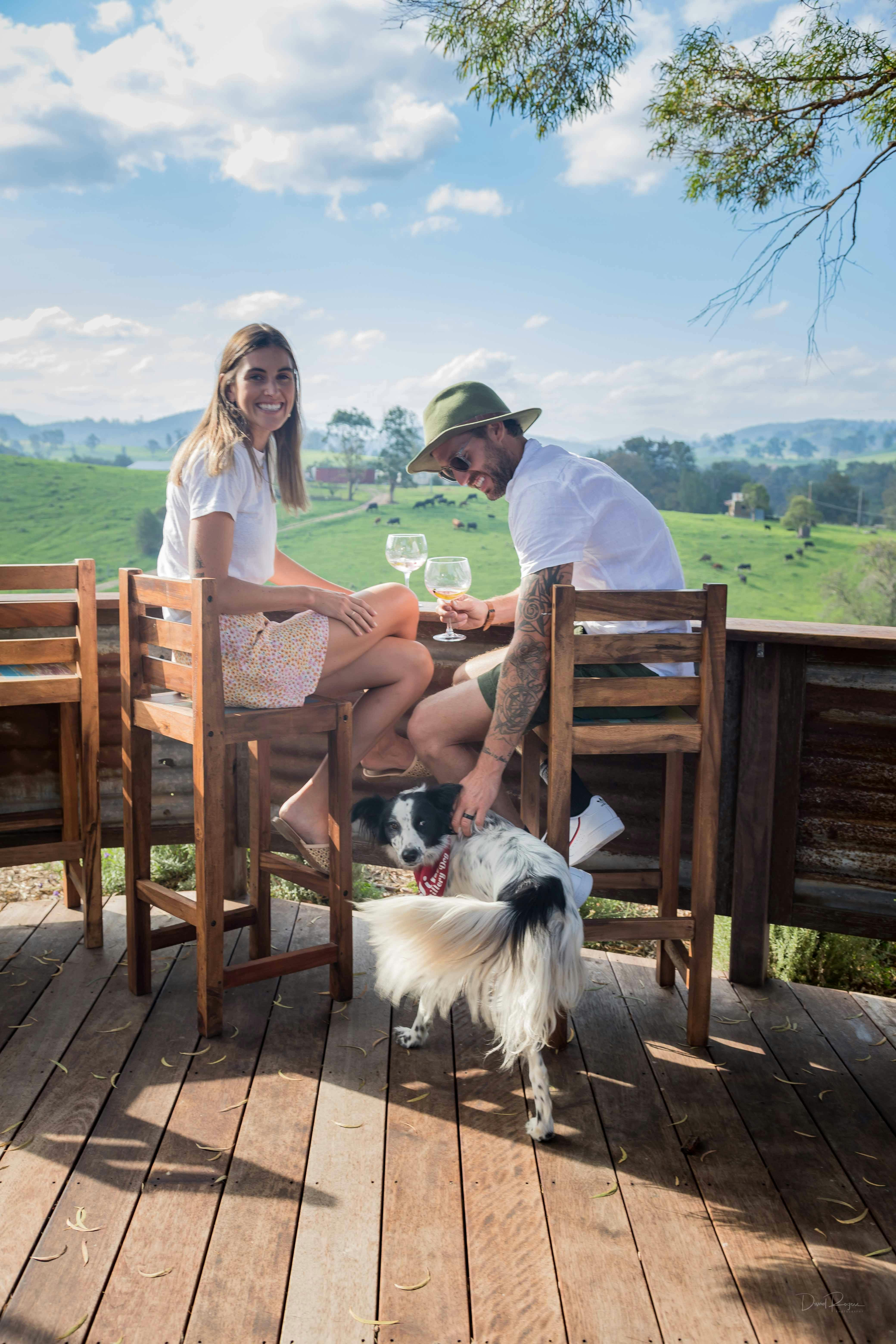 Young man and woman sitting on a deck with drinks and a dog