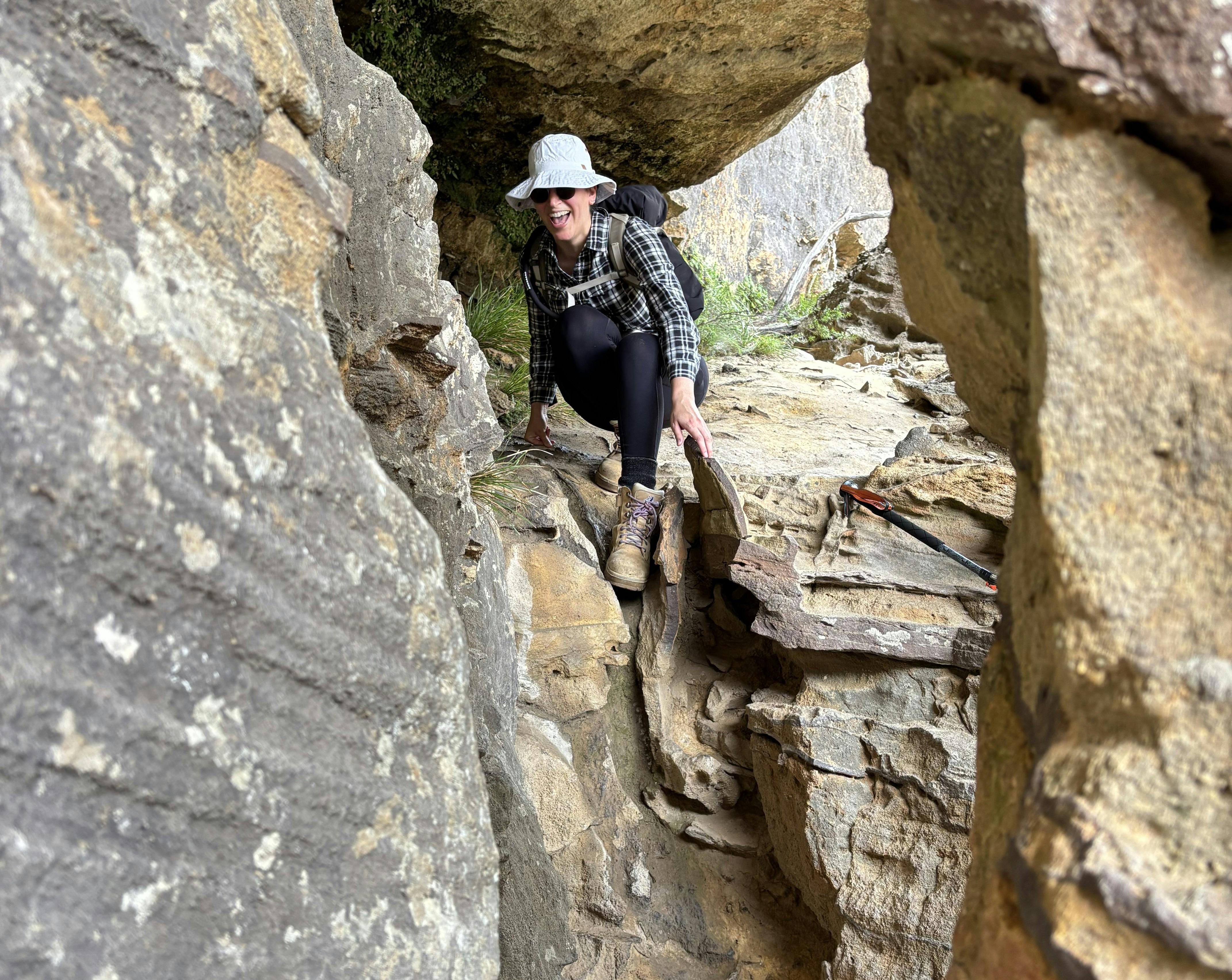 A woman wearing a checked shirt and black trousers climbing down a sand stone ledge