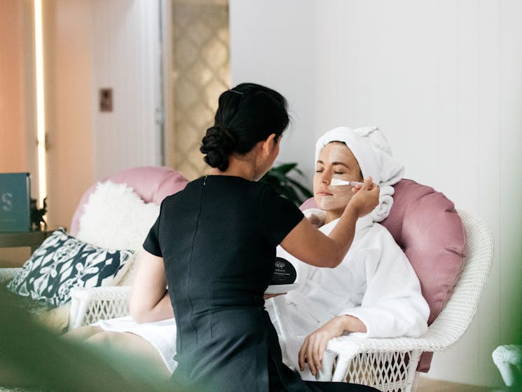 A therapist is spreading face mask onto a woman in a robe lying in a plush pink lounge chair