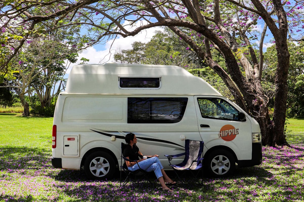 A woman sits in a campchair under the shade of a jacaranda tree next to a Hippie Endeavour Camper