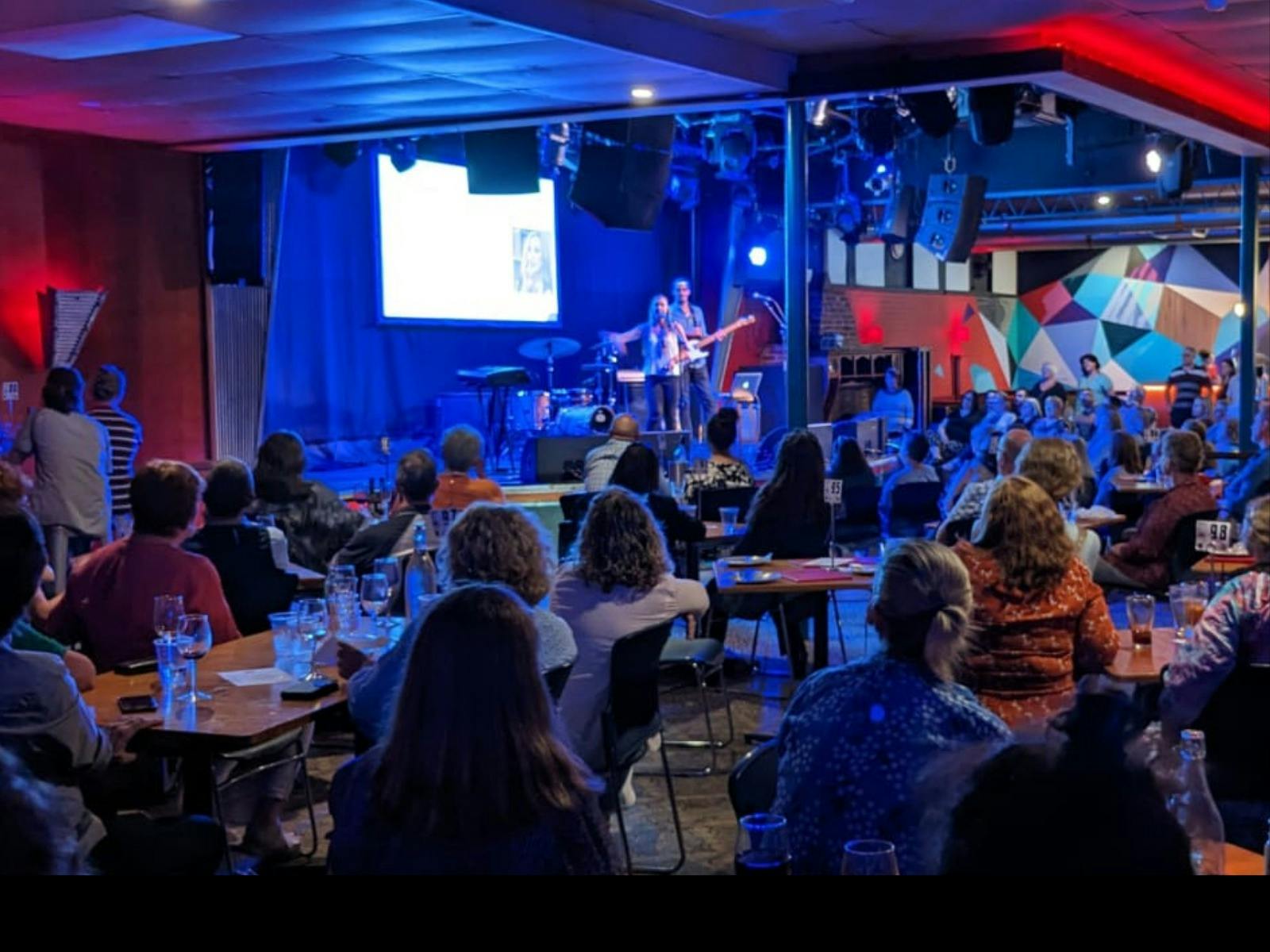 A crowd watches a musician perform on a small stage in a lively club, with blue and red lights