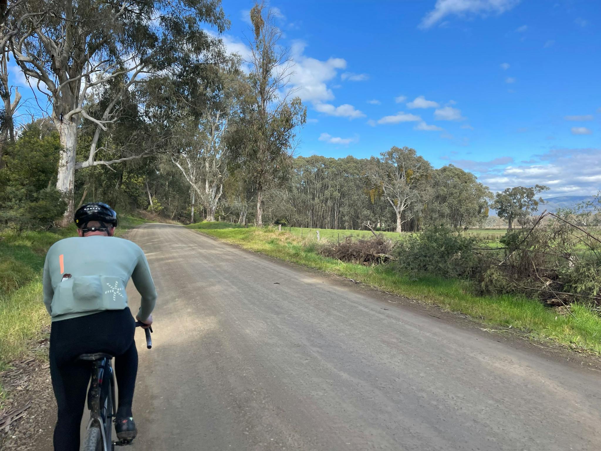cyclist on side of gravel road, bushy trees on right green grass, gum trees, blue sky and clouds