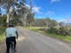 cyclist on side of gravel road, bushy trees on right green grass, gum trees, blue sky and clouds