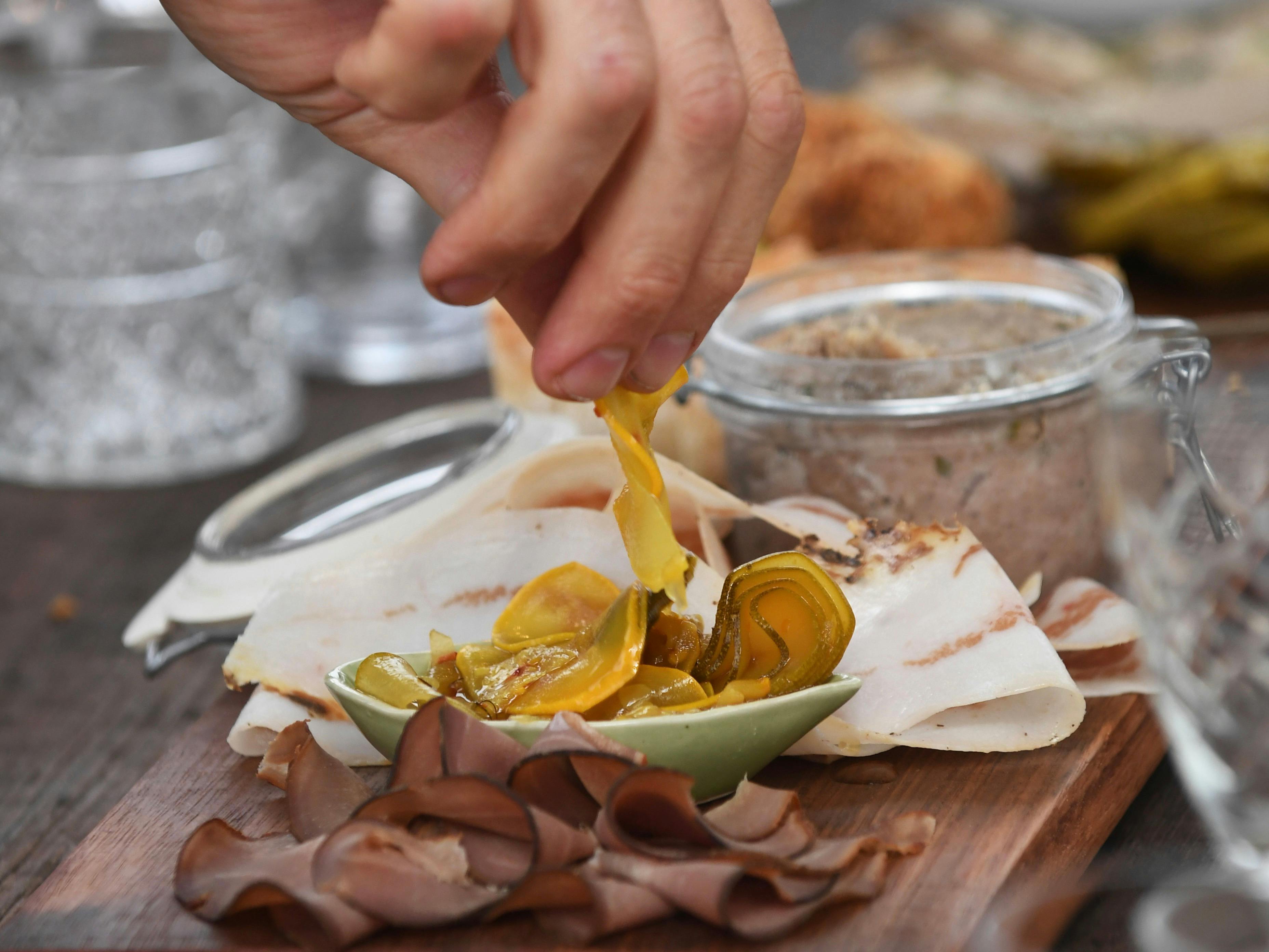Hand picking charcuterie from a platter with sliced meats, pickles, and spreads on a wooden board.