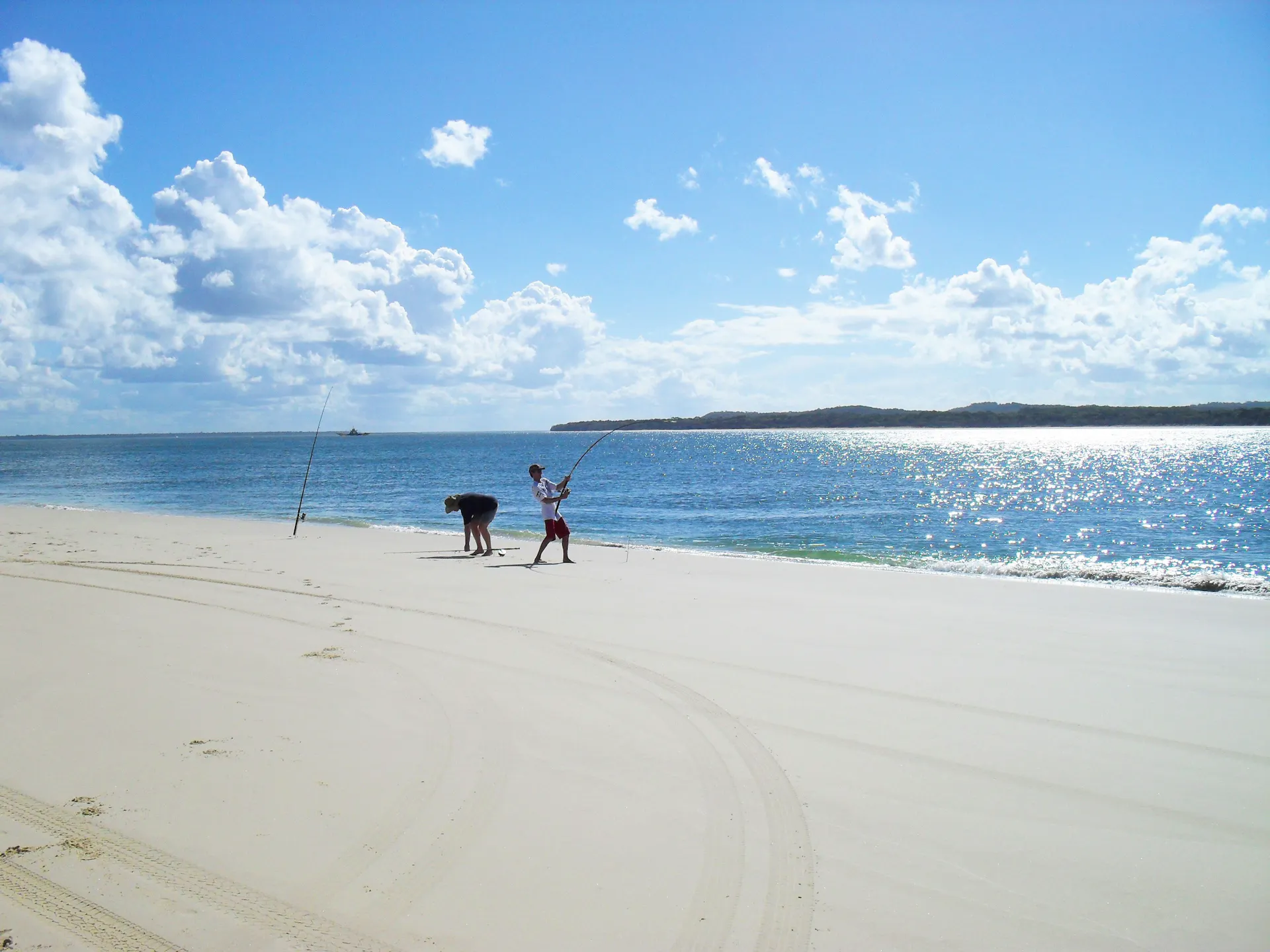 Two people line fishing at waters edge on sandy beach with blue skies overhead.