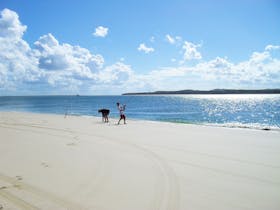 Two people line fishing at waters edge on sandy beach with blue skies overhead.