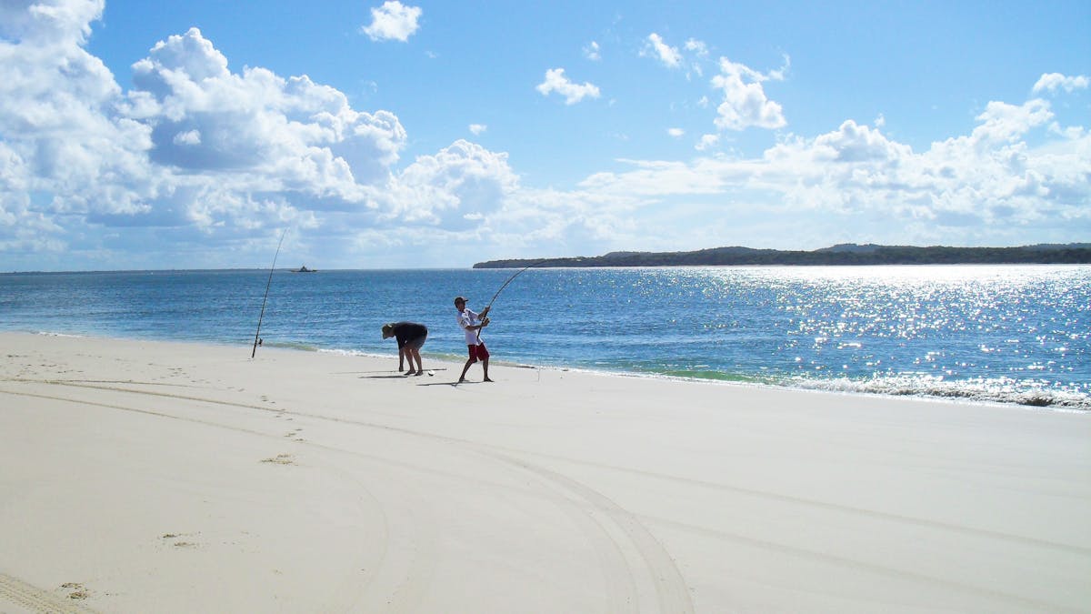 Two people line fishing at waters edge on sandy beach with blue skies overhead.