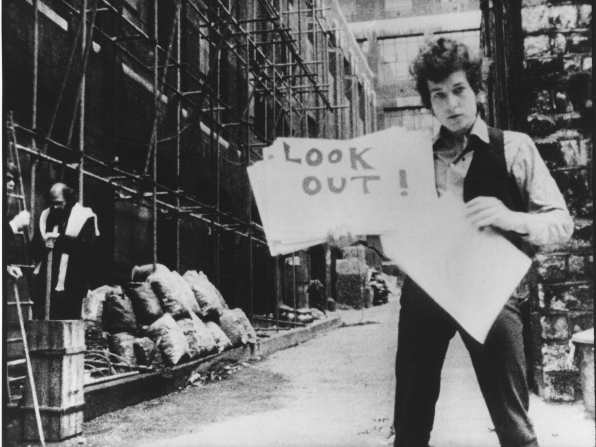 Black and white photo of Bob Dylan holding a poster that says look out!