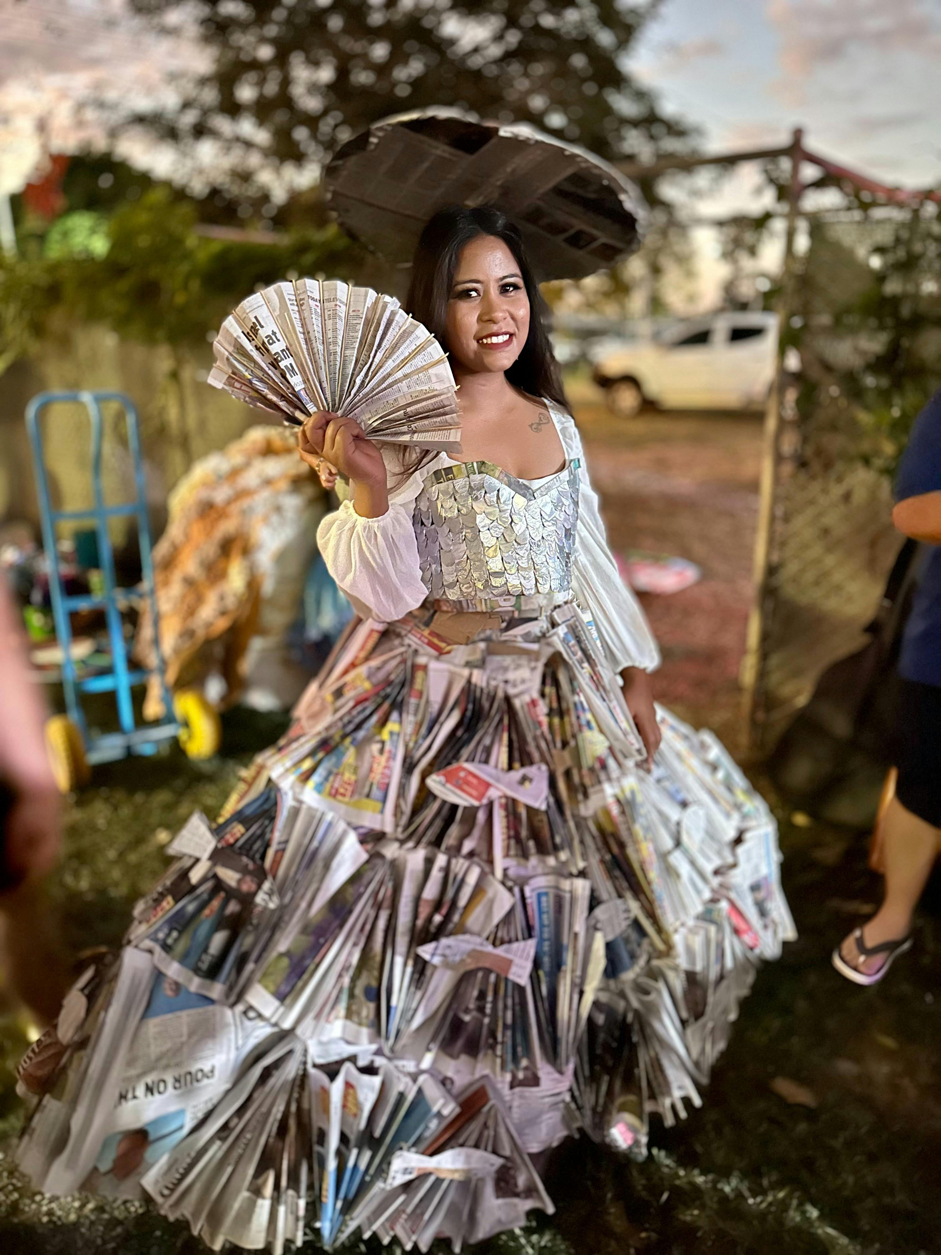 A woman wears a long dress and holds a fan all made out of newspaper for the junk fashion parade