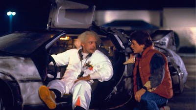 Two men facing each other, man on left sitting in car in white doctors suit