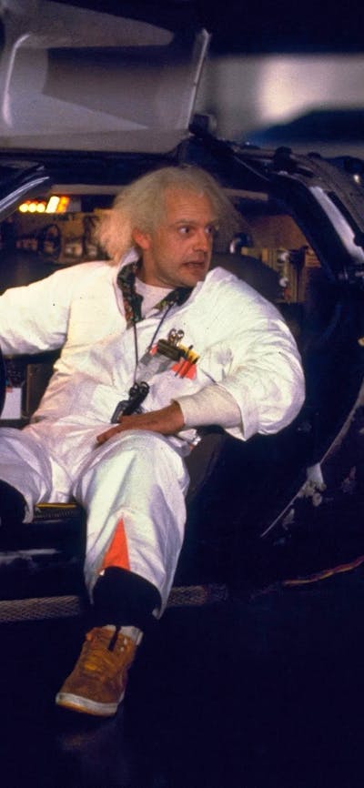 Two men facing each other, man on left sitting in car in white doctors suit