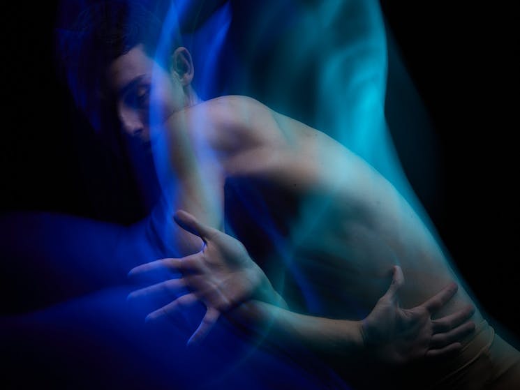 One dancer bathed in blue light crosses over their arms with their left palm outstretched.
