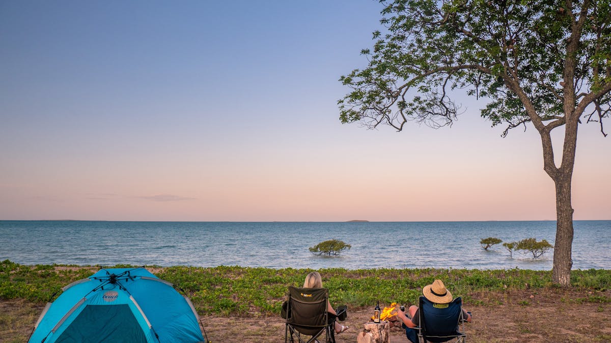 Blue tent with 2 people sitting in chairs watching the sunset camped under a tree.