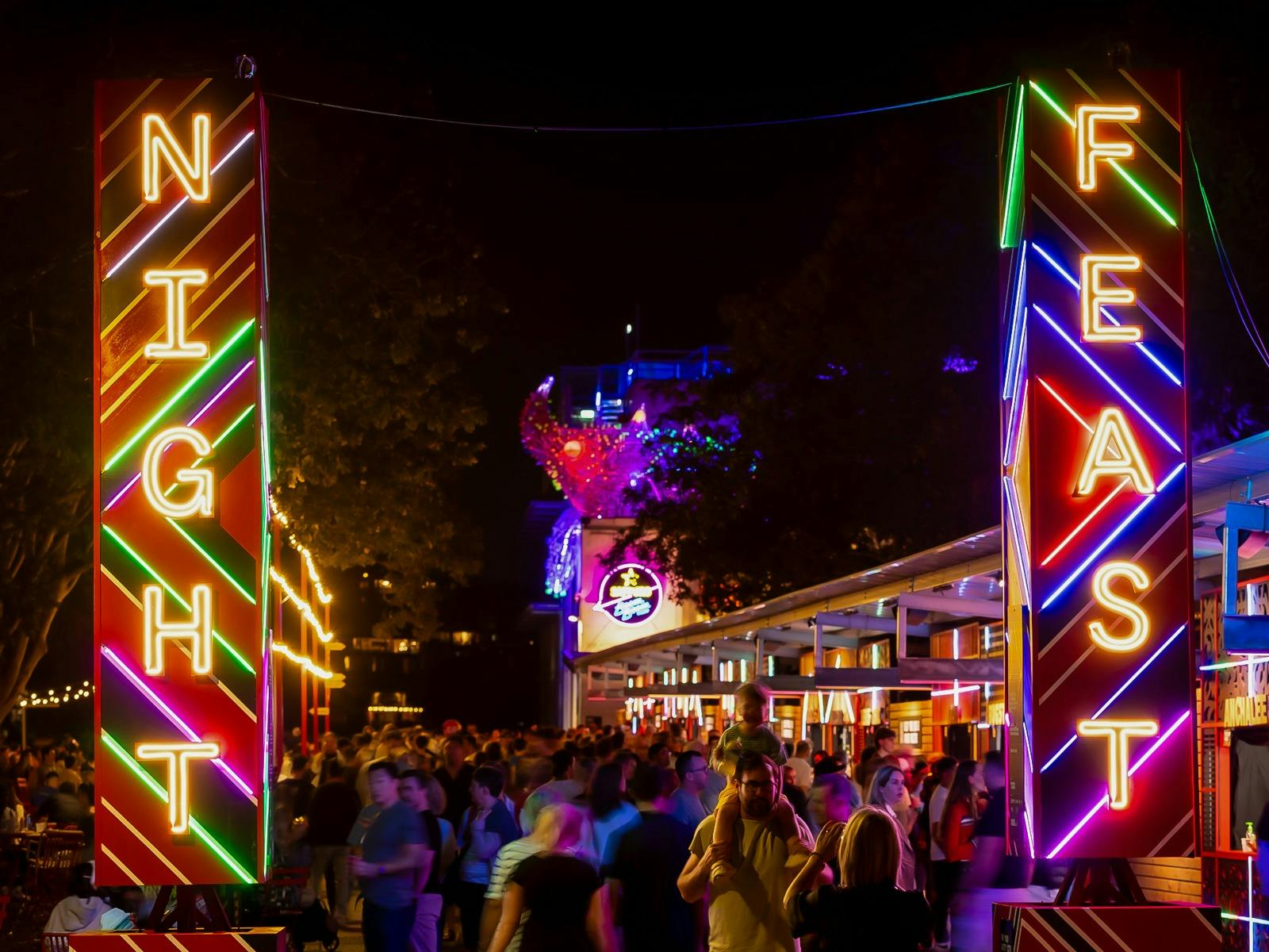 Neon “Night Feast” signs glow over a bustling outdoor event with crowds, color, and festive energy. Neon “Night Feast” signs glow over a bustling outdoor event with crowds, color, and festive energy.