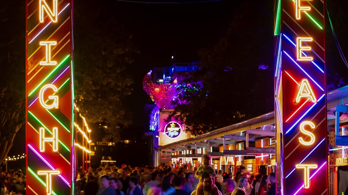 Neon “Night Feast” signs glow over a bustling outdoor event with crowds, color, and festive energy.