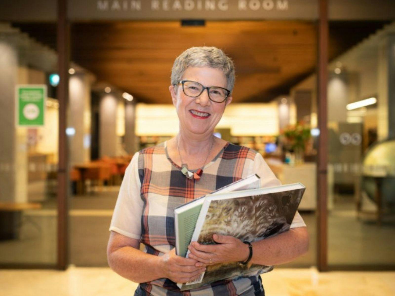 A smiling woman wearing glasses is pictured in a Library holding books