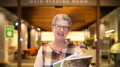 A smiling woman wearing glasses is pictured in a Library holding books