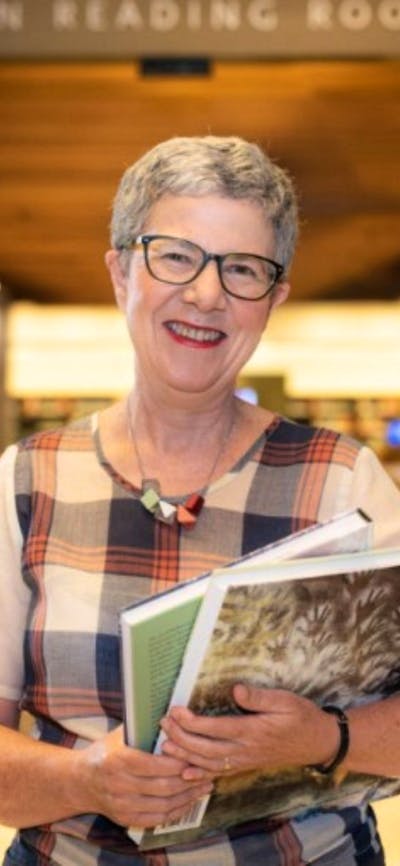 A smiling woman wearing glasses is pictured in a Library holding books