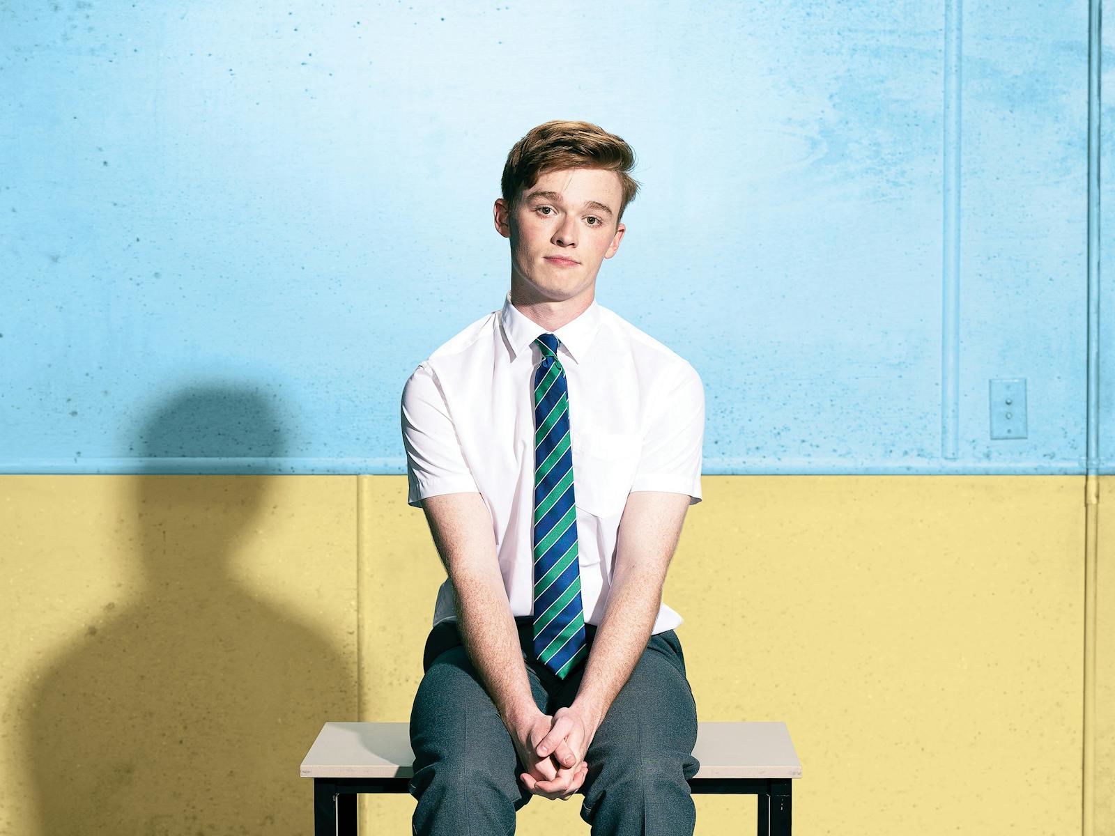 A young man in school uniform, sits on a school desk, looking at the camera