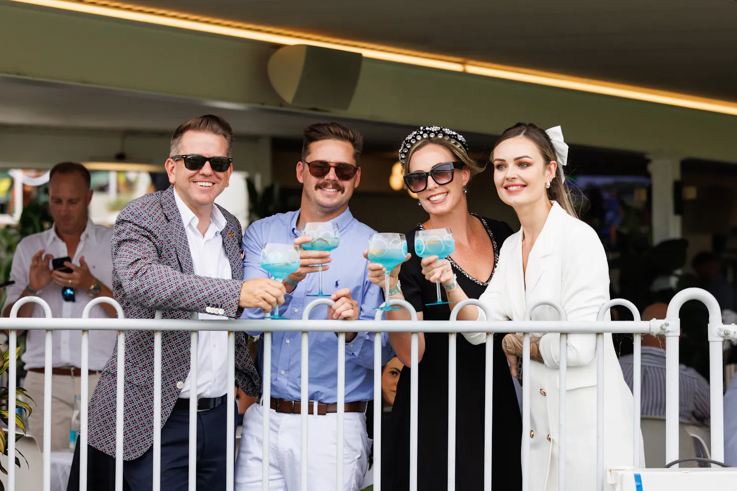 Four people on the fence line holding up their cocktail glasses and smiling at the camera